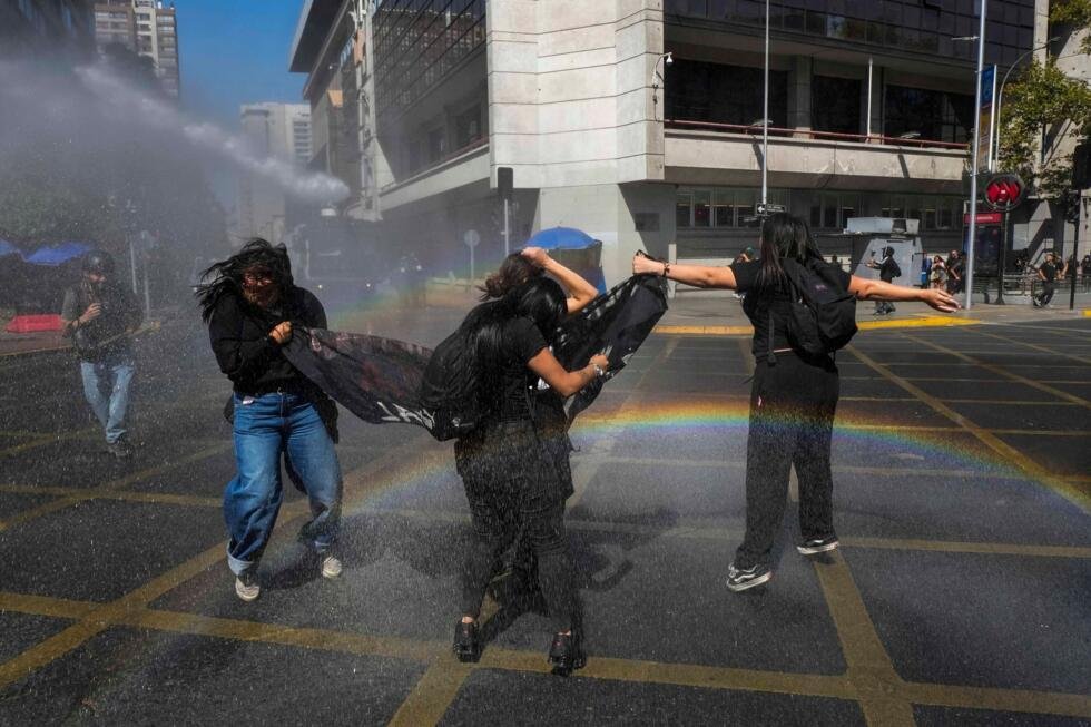 Jóvenes se preparan para un cañón de agua policial durante una protesta estudiantil contra la propuesta del gobierno de reducir el presupuesto educativo y contra el aumento del precio de la gasolina en Santiago de Chile, el jueves 26 de marzo de 2026. (Foto AP/Esteban Felix)