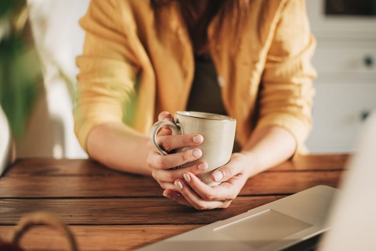 Por la mañana, café para el desayuno y manos femeninas vertiendo agua en una taza den la cocina de casa. Mujer en pijama preparando té o alguna bebida en la encimera para empezar el día.