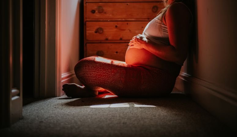 Una futura madre descansa apoyada contra una pared en un pasillo estrecho y poco iluminado, acariciando su vientre mientras disfruta de un momento de tranquilidad y reflexión al caer la tarde.