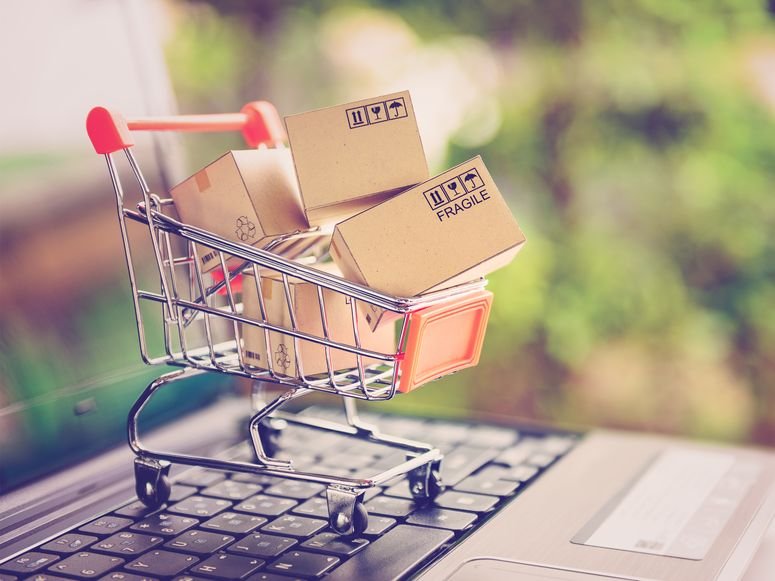 Carrito de compra en miniatura con pequeñas cajas de cartón en su interior, sobre las teclas de una laptop.